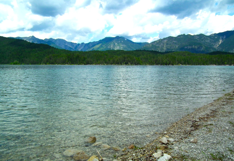 klarer Bergsee mit Kieselstrand und bewaldeten Bergen unter wolkigem Himmel