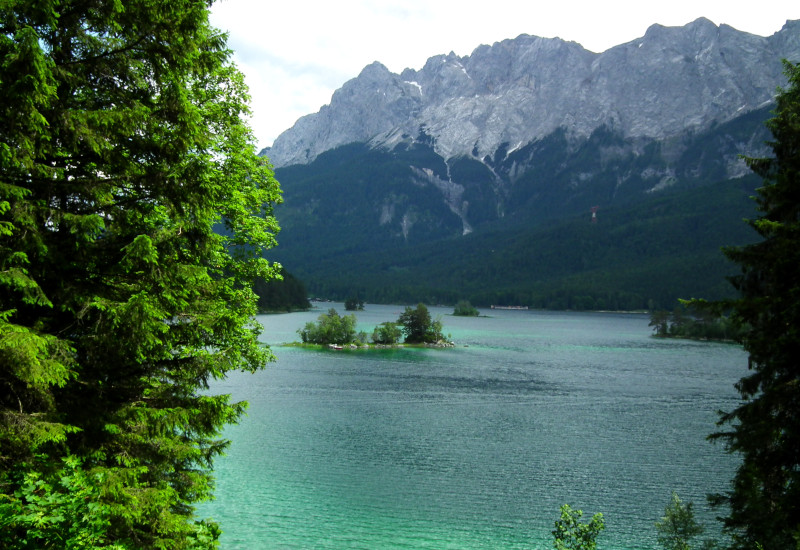 Blick auf einen klaren Bergsee (Eibsee) mit grüner Insel und bewaldeten Ufern, dahinter hohe, felsige Berge