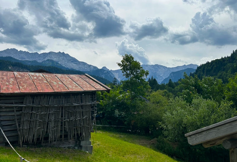 Altes Holzhaus mit rostigem Blechdach vor grünen Bäumen und Bergpanorama unter bewölktem Himmel