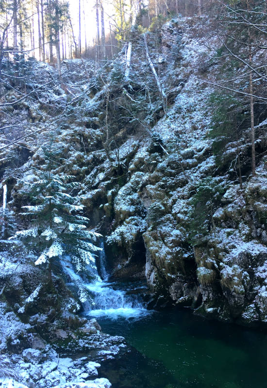 Bach mit Wasserfall in verschneiter, bewaldeter Landschaft.
