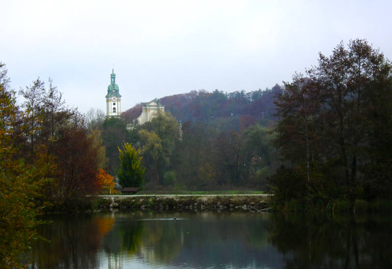 Herbstliche Landschaft mit Teich und Kirche im Hintergrund.