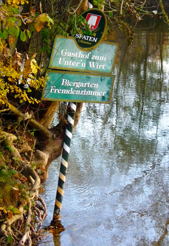 Schild mit Gaststätteninformationen am Wasser, umgeben von Bäumen.