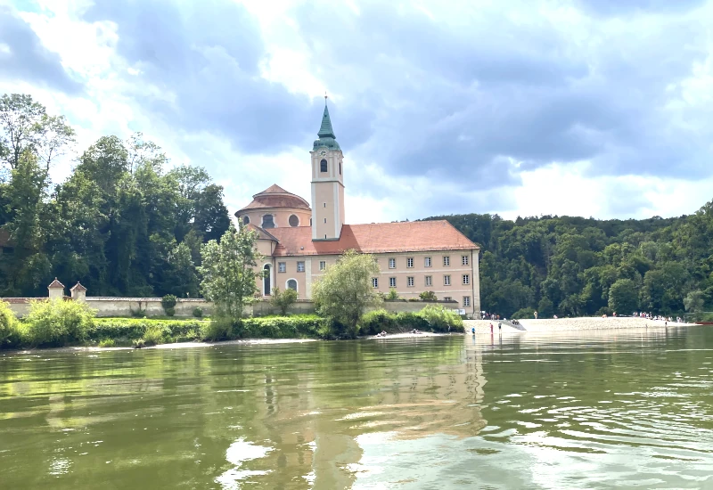 Schloss am Fluss mit bewaldetem Hintergrund und bewölktem Himmel.