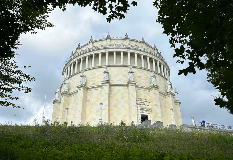 Monumentaler Rundbau mit Säulen und Statuen, umgeben von Bäumen.