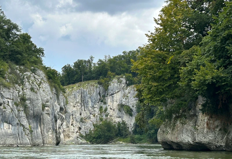 Felsen und Bäume entlang eines ruhigen Flusses unter bewölktem Himmel.