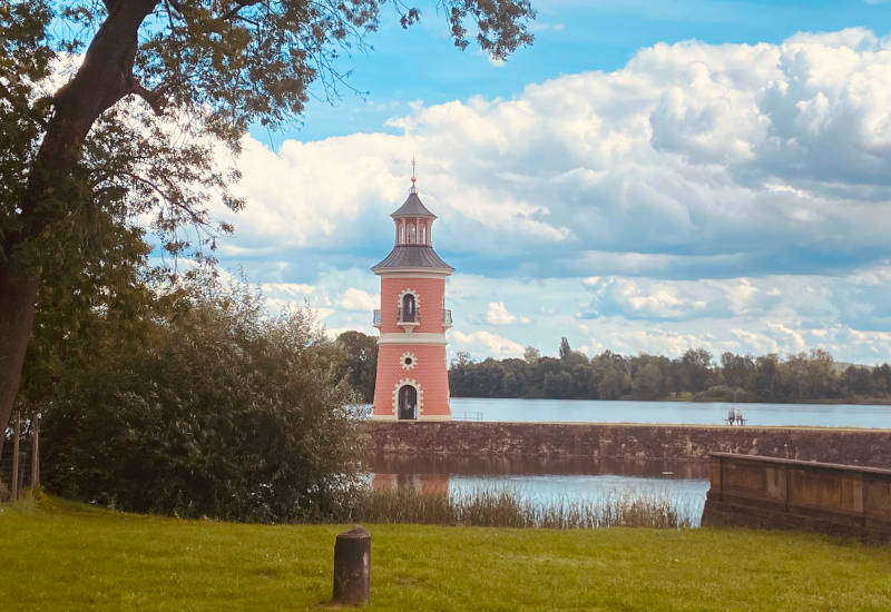 Roter Wasserturm am Ufer eines Sees unter bewölktem Himmel.