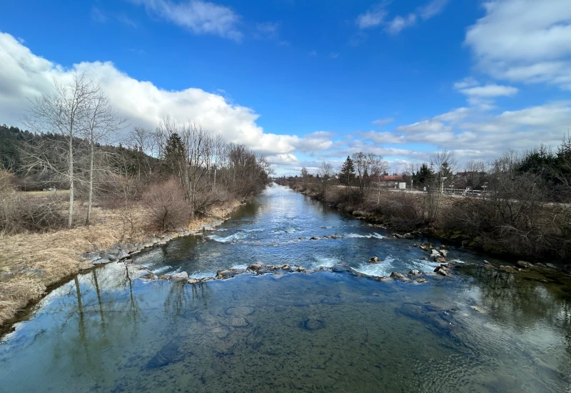 Fluss mit klarem Wasser, umgeben von Bäumen und Wolken.