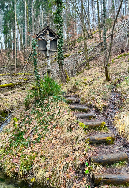 Holzkreuz an einem Waldweg mit Stufen und Laub.